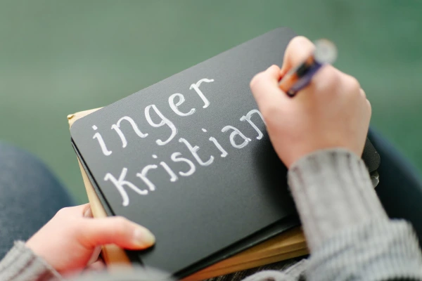 Close-up of hands writing the names "Inger Kristian" in white chalk lettering on a dark notebook cover. Candid wedding detail shot.