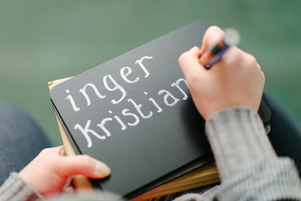 Close-up of hands writing the names "Inger Kristian" in white chalk lettering on a dark notebook cover. Candid wedding detail shot.
