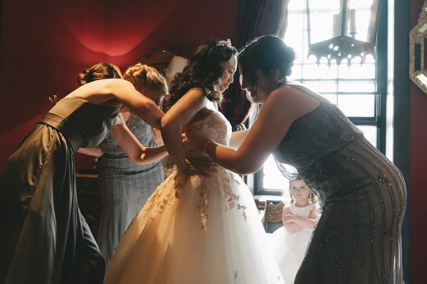 Bride in white lace and tulle surrounded by supportive women, warm golden light through the window
