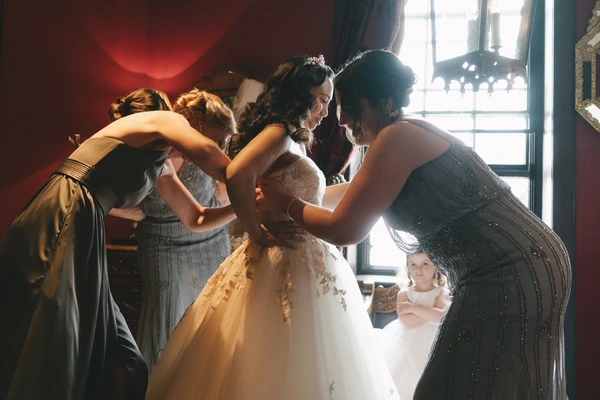 Bride in white lace and tulle surrounded by supportive women, warm golden light through the window