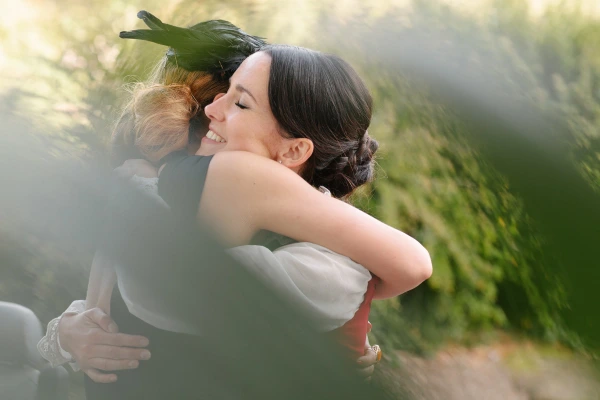 The bride laughs joyfully with closed eyes as she embraces a guest with a feathered fascinator outdoors in warm golden light. Candid documentary wedding portrait.