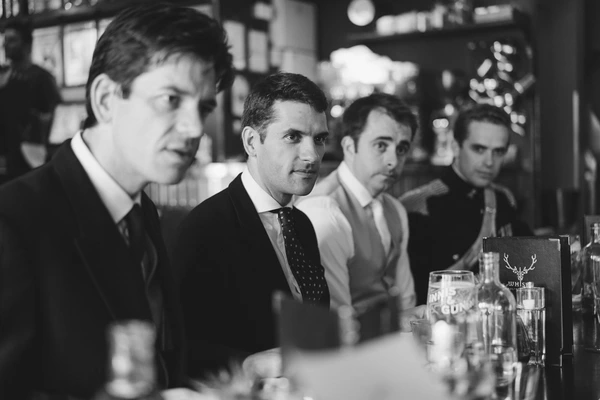 Four suited men at the bar, serious expressions — black-and-white, whisky and glasses in foreground