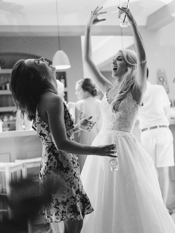 Bride in white dances joyfully with a female guest — both arms raised, natural light, candid moment