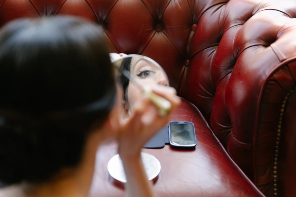 Bride applying mascara using a small round compact mirror, seated against a red Chesterfield sofa during morning preparations. Candid getting-ready moment.