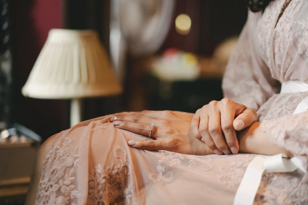 Bride's ring-bearing hand rests on the groom's arm in lace — warm lamp light on the jewel
