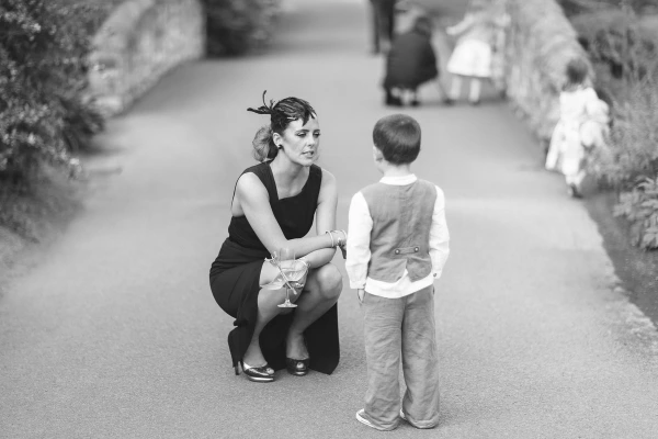 Black and white image of a woman in a black dress and feathered fascinator crouching to speak to a young boy on a garden path, with more children playing in the background. Candid documentary wedding moment.