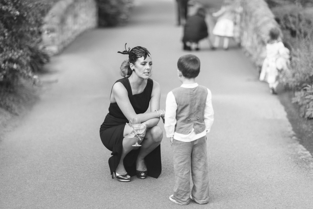 Black and white image of a woman in a black dress and feathered fascinator crouching to speak to a young boy on a garden path, with more children playing in the background. Candid documentary wedding moment.