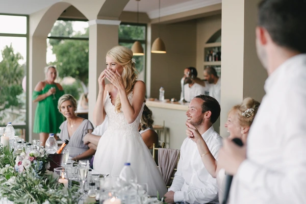 Bride covers her mouth in delighted surprise, groom looks on astonished — outdoor reception