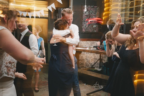 Father holds the little boy at a reception amid lights — guests dancing, decorated bar beside them