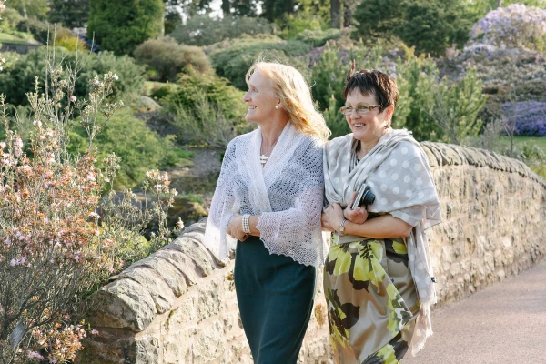 Two elegantly dressed women walk arm in arm along a stone-walled garden path at the Royal Botanic Garden Edinburgh, smiling and chatting. Candid documentary wedding photograph.