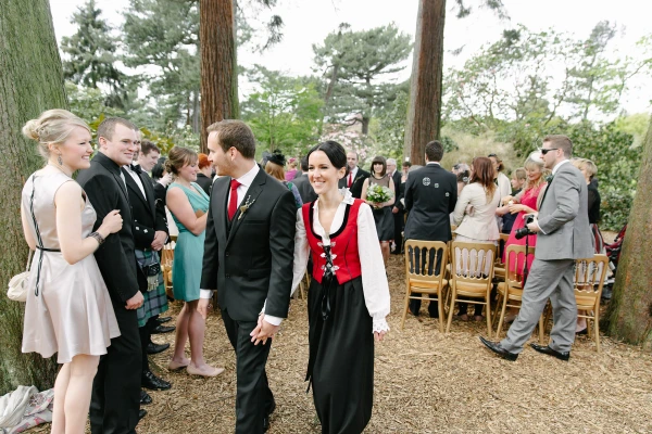 Couple walking hand in hand down the outdoor ceremony aisle as guests and guests in kilts applaud around them. Candid post-ceremony recessional.