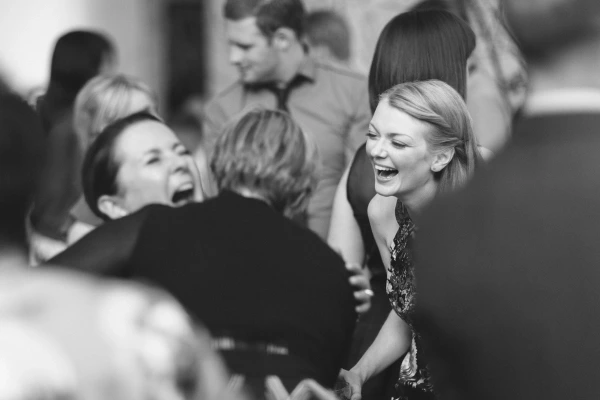 Black and white candid image of wedding guests laughing together on the dance floor, a blonde woman in the foreground beaming with delight. Documentary wedding photography.