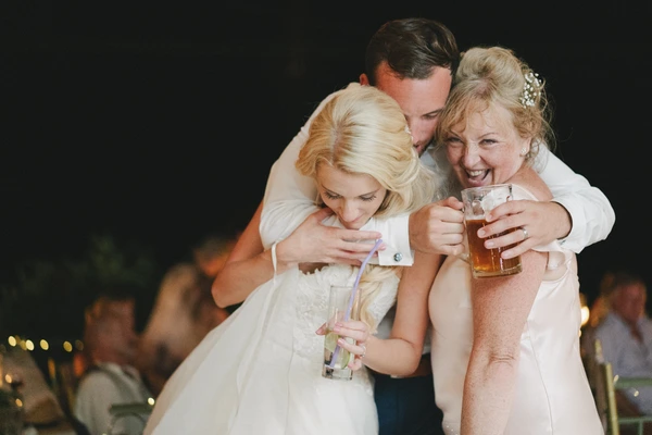 Groom embraces smiling bride and parent at the celebration — glasses in hand, evening party