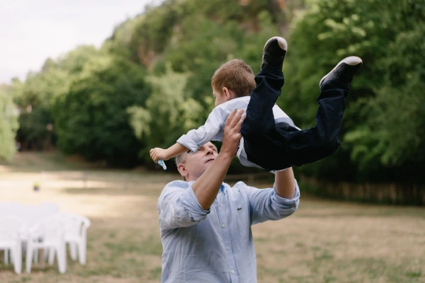 Father lifting young son in air playfully with lush green valley landscape background at wedding.