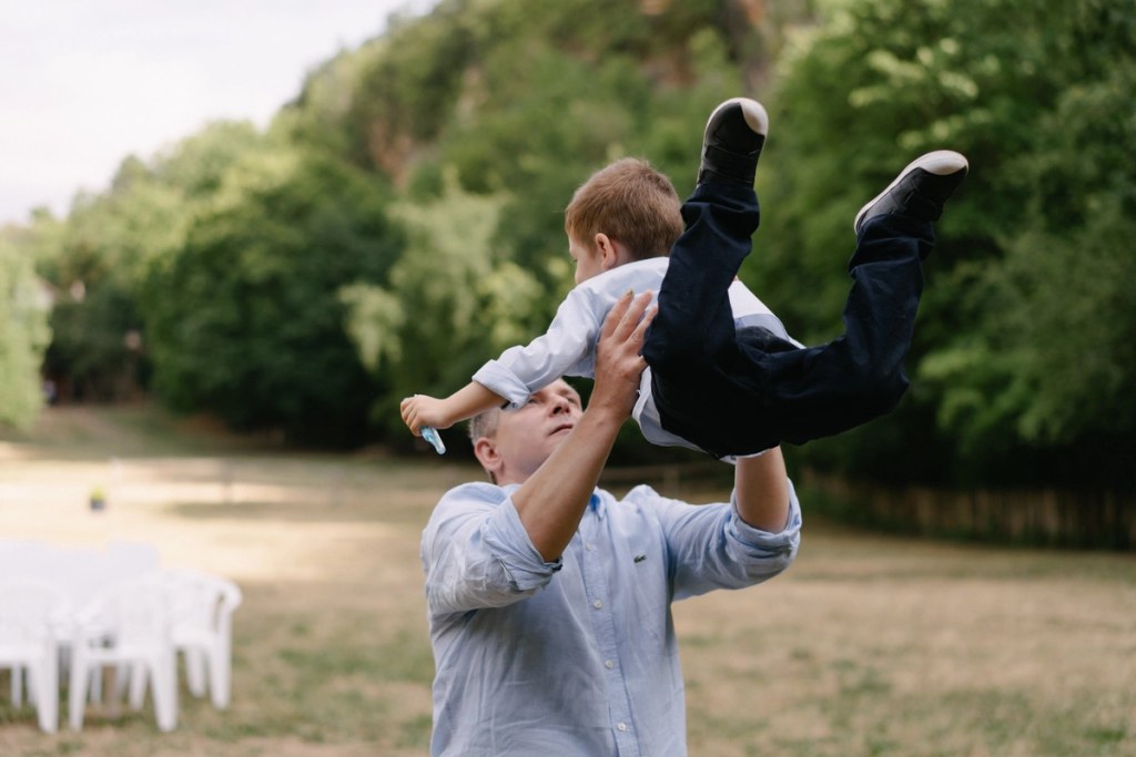 Father lifting young son in air playfully with lush green valley landscape background at wedding.
