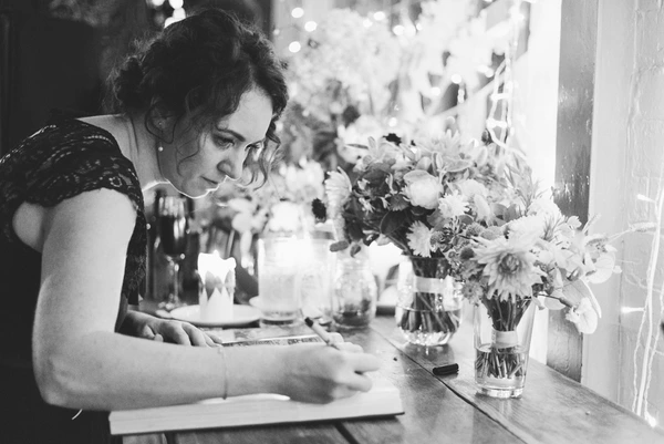 Woman in black lace writes at a table, fresh flowers in glass blocks, natural window light