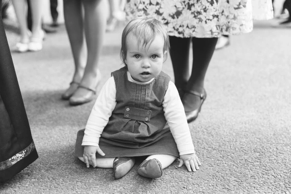 Black and white candid portrait of a baby girl in a pinafore dress sitting on the ground at the wedding, looking directly into the camera. Charming documentary moment.