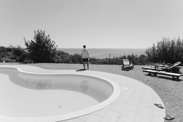 Man in white polo stands by the pool looking out to sea — curved pool, pergola, black-and-white