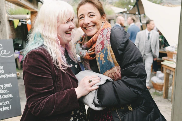 Two women laughing and talking among wedding guests — warm embrace, joyful party garden