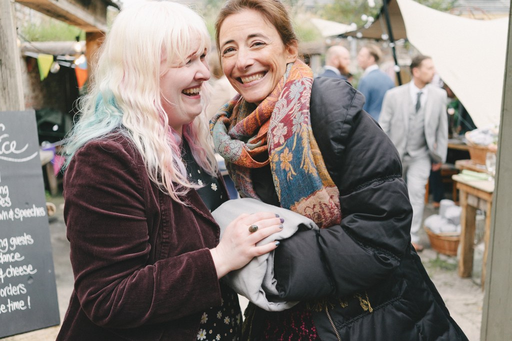 Two women laughing and talking among wedding guests — warm embrace, joyful party garden