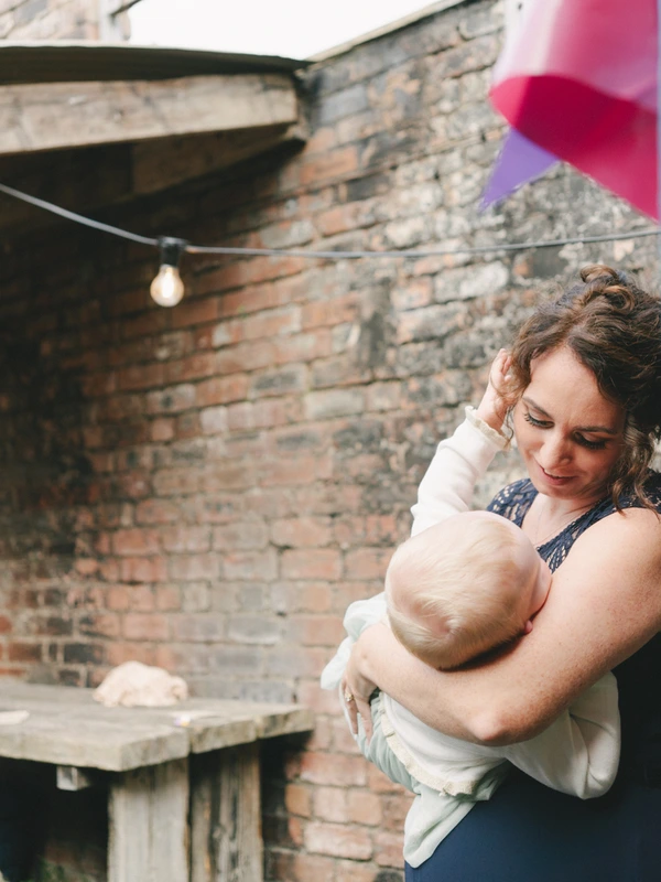 Mother tenderly holds her baby in a courtyard — pink balloon in background, warm sunlight