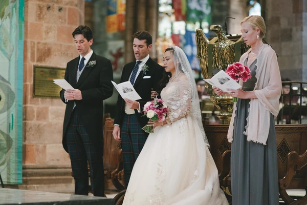 Bride in white lace at the altar with bouquet, groom in tartan kilt, witnesses reading