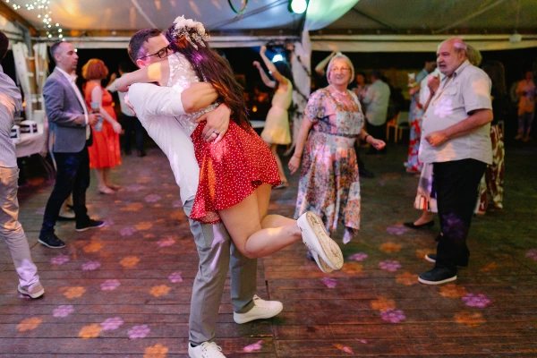 Groom lifts bride playfully on dance floor — colorful ambient lighting, romantic celebration, joyful couple moment at reception.