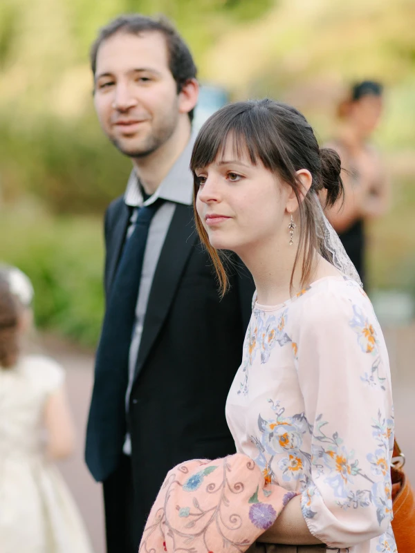 A young couple stands together among other guests at a garden wedding, the woman in a floral dress looking quietly off to the side. Candid documentary wedding photograph.