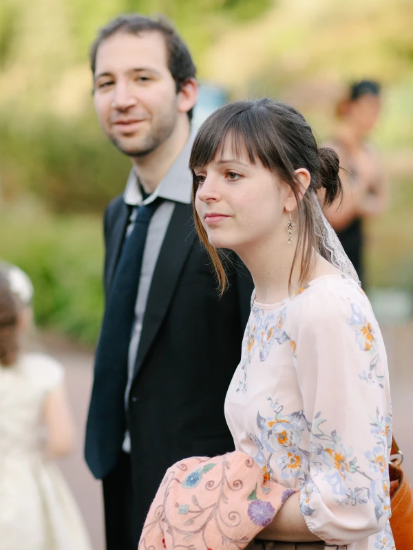 A young couple stands together among other guests at a garden wedding, the woman in a floral dress looking quietly off to the side. Candid documentary wedding photograph.