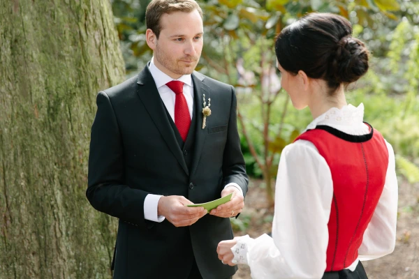 Black and white candid portrait of the couple leaning toward each other during the ceremony as the officiant reads from a folder beside them. Intimate outdoor ceremony moment.