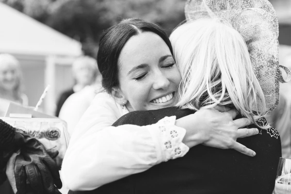 Black and white candid close-up of the bride smiling with her eyes closed as she hugs a blonde guest in a lace fascinator warmly after the ceremony. Emotional embrace.