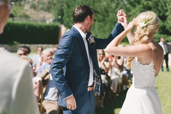 Bride in white lace dances with groom in dark blue suit — guests watching in the sunny garden