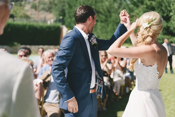 Bride in white lace dances with groom in dark blue suit — guests watching in the sunny garden