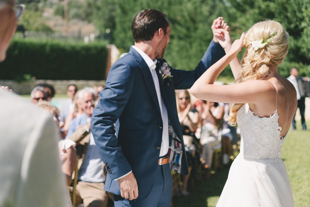 Bride in white lace dances with groom in dark blue suit — guests watching in the sunny garden