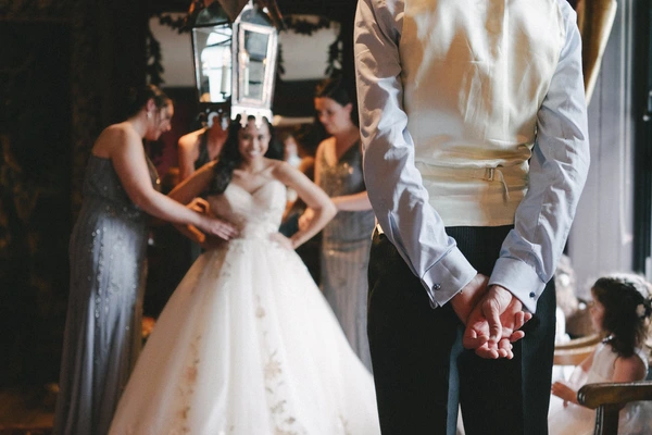 Groom in white shirt and black suit holds the bride's hand at the ceremony, white lace gown behind