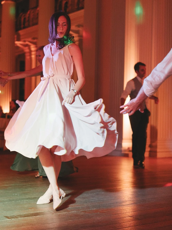 Young woman in white spins dancing in a red-lit hall, floral necklace, partner holds her hand