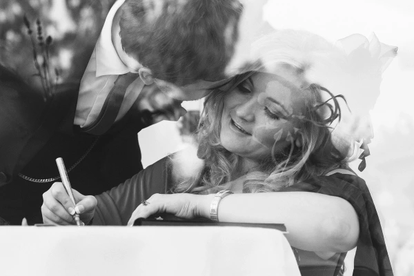 Black and white candid shot of a male guest leaning over to kiss a laughing female guest who is signing the guestbook at the reception. Playful and affectionate moment.