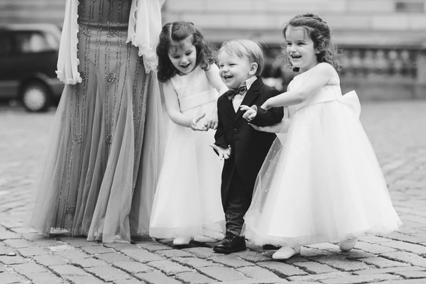 Three cheerful little girls and a boy in wedding attire play on cobblestones, holding the bride's train