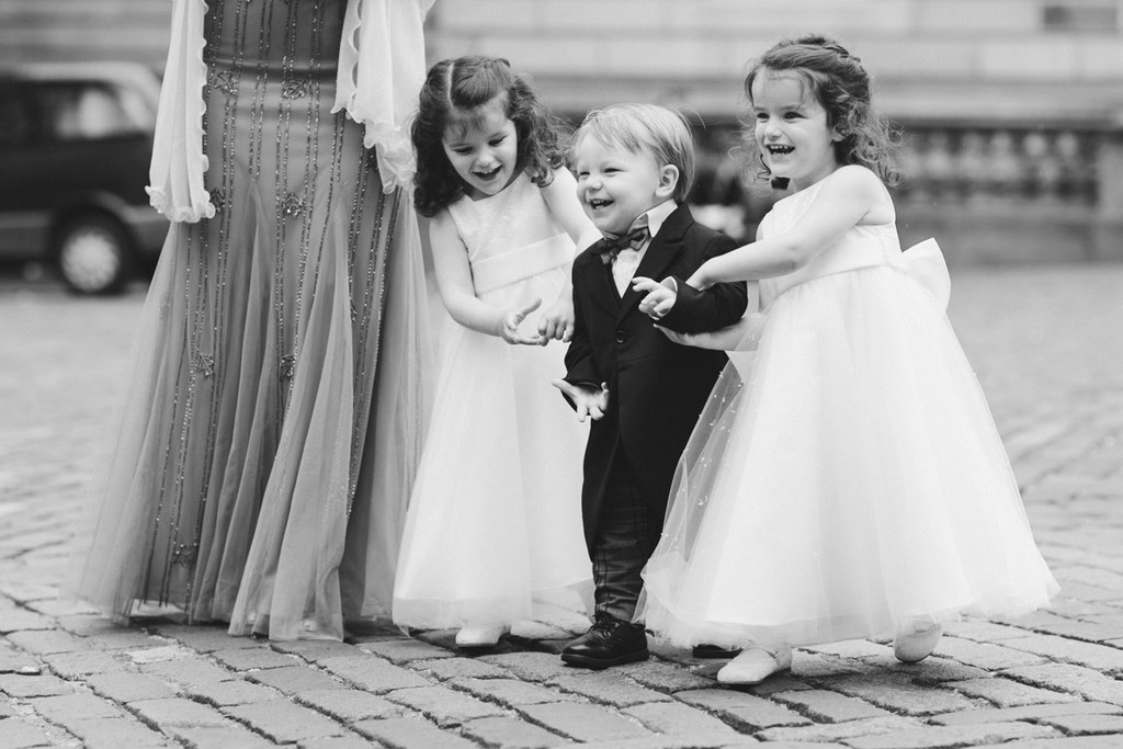 Three cheerful little girls and a boy in wedding attire play on cobblestones, holding the bride's train