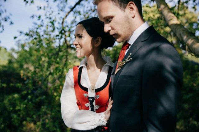 Smiling bride embraces groom in dark suit outdoors among green trees on sunny wedding day