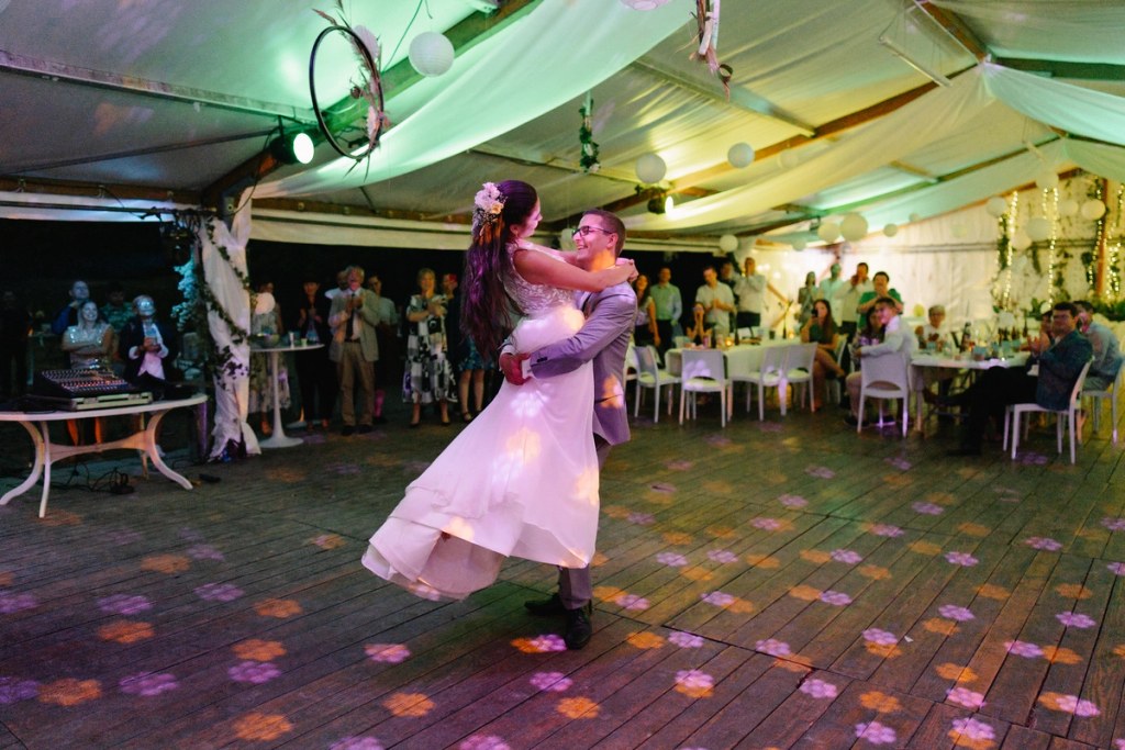 Bride and groom dancing at evening wedding reception with guests watching under tent lights.