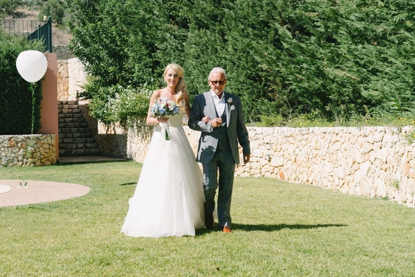 Bride arm in arm with father walking through the courtyard, bouquet in hand, stone walls around