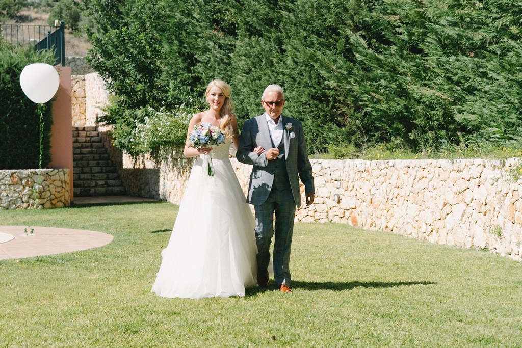 Bride arm in arm with father walking through the courtyard, bouquet in hand, stone walls around