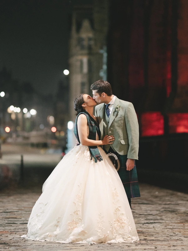Newlyweds kiss on a city street at night — white lace, tartan suit, lit buildings behind