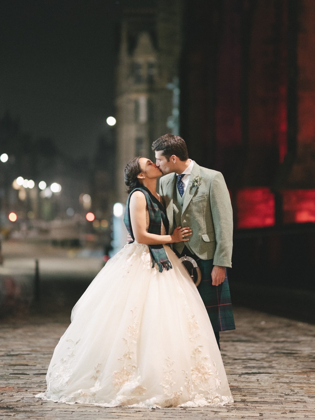 Newlyweds kiss on a city street at night — white lace, tartan suit, lit buildings behind