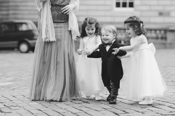 Elegant woman with pearl scarf surrounded by three cheerful children in wedding attire on cobblestones