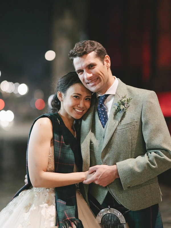 Young couple smiling in night city light — groom in green jacket and tartan, bride in white lace