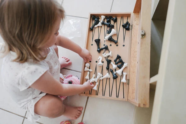 Little girl crouches choosing a chess piece from a wooden box — concentrated, light floor and wall