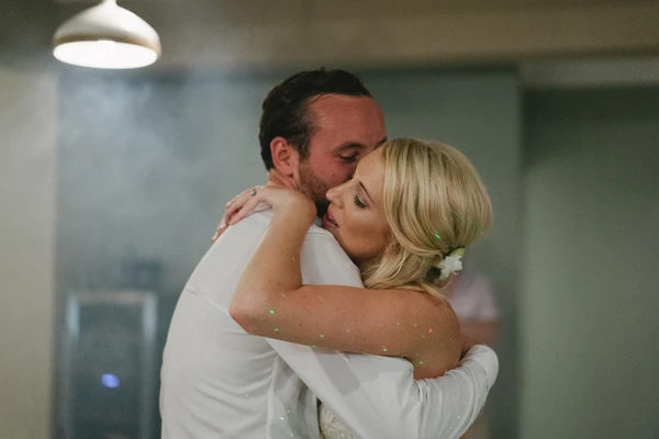 Groom in white shirt kisses bride — she wears a white gown and sparkle-decorated arm, warm light