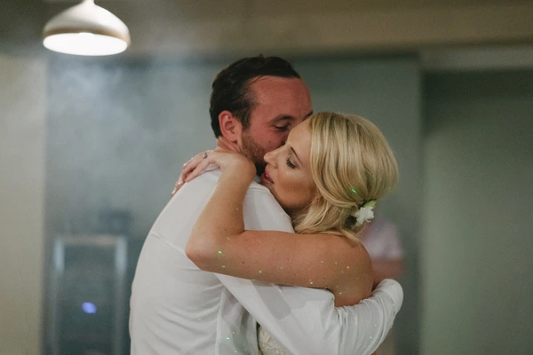 Groom in white shirt kisses bride — she wears a white gown and sparkle-decorated arm, warm light